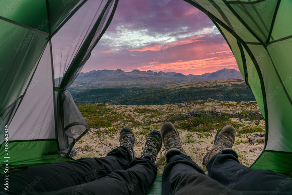 Couple with hiking boots are laying with feet outside the tent opening with a beautiful view over the mountains in the back with amazing sunrise red colored skies. Camping and landscape concept.