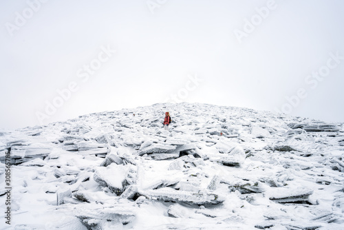 Girl with red hooded jacket and backpack are hiking in snow covered mountains with foggy weather. Scenery, lifestyle, active and outdoor concept.