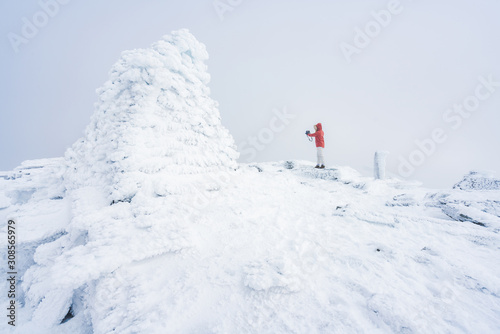 Girl with red hooded jacket and hoodie are taking a selfie next to a frozen cairn on top of a mountain peak covered with snow and fog. Scenery, lifestyle, active and outdoor concept. 