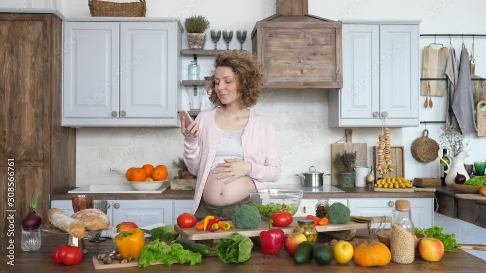 Beautiful Pregnant Female Using Smartphone And Cooking On Kitchen. Beautiful Pregnant Female Using Smartphone And Cooking On Kitchen.