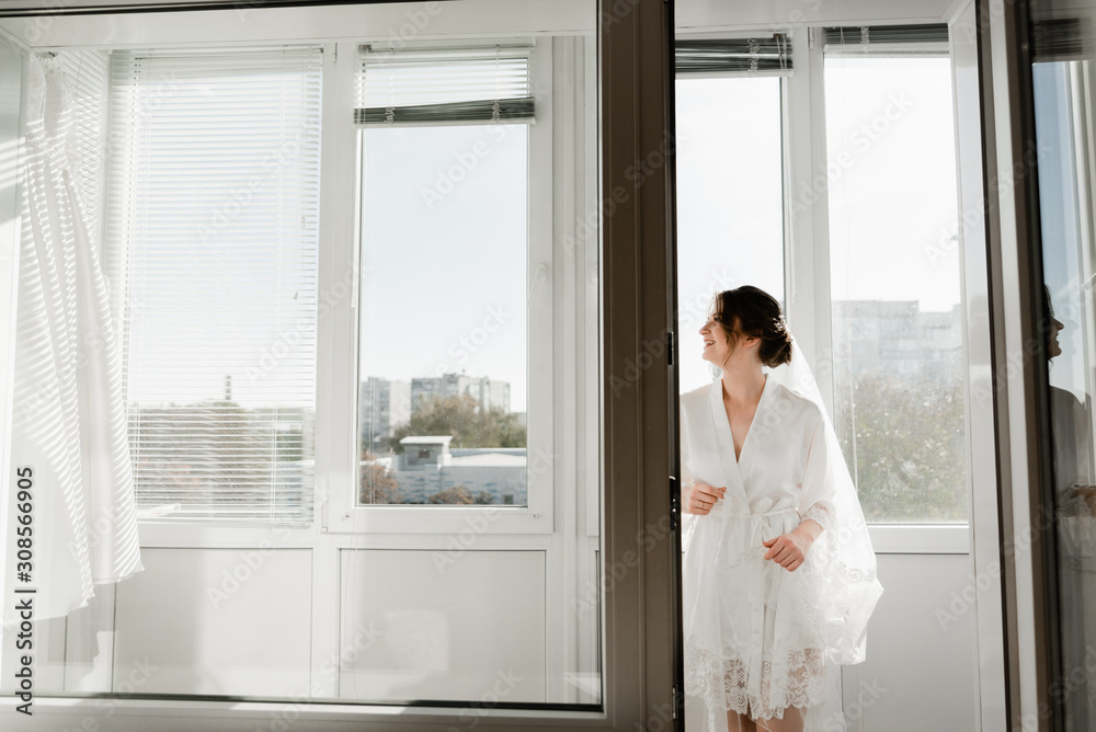 Portrait of young gorgeous bride on the balcony. Beautiful girl dresses and gathers for a wedding ceremony in apartments while waiting for the groom. Bride looking at her dress. Bride's morning Portrait of young gorgeous bride on the balcony. Beautiful girl dresses and gathers for a wedding ceremony in apartments while waiting for the groom. Bride looking at her dress. Bride's morning