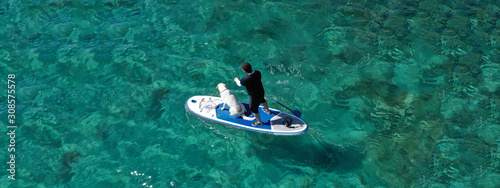 Aerial drone ultra wide panoramic photo of unidentified fit man paddling with his cute dog on a SUP board or Stand Up Paddle board in tropical exotic emerald bay