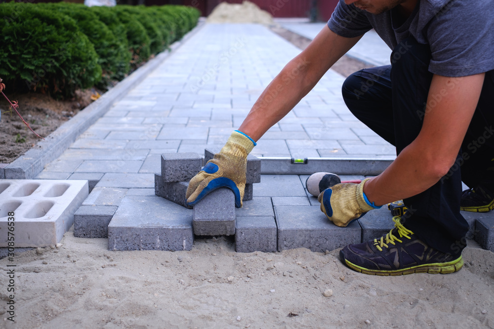 The master in yellow gloves lays paving stones in layers. Garden brick pathway paving by professional paver worker. Laying gray concrete paving slabs in house courtyard on sand foundation base. The master in yellow gloves lays paving stones in layers. Garden brick pathway paving by professional paver worker. Laying gray concrete paving slabs in house courtyard on sand foundation base.