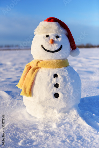Funny snowman in red santa hat and yellow scalf on snowy field. Christmass and New Year background
