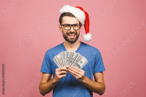 Happy Bearded man in casual and christmas hat holding money and looking at the camera isolated over pink background. photo