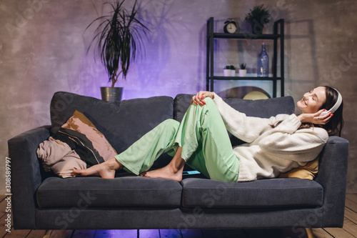 A young woman is lying on the couch in a cozy room listening to music on wireless headphones. Girl happy. photo