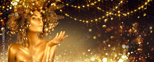 Christmas golden Woman. Winter girl pointing Hand, blowing blinking stars, Beautiful New Year, Christmas Tree Holiday Hairstyle and gold skin Makeup. Gift. Girl in decorated Xmas wreath. Beauty Model