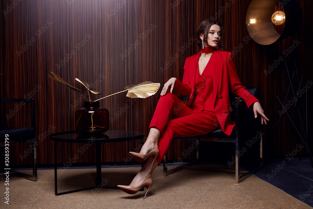 Beautiful young pretty woman bright evening makeup of shiny red lipstick brunette curly hair wear elegant suit red trousers blouse jacket high heels clothes for party meeting interior room fashion. St...