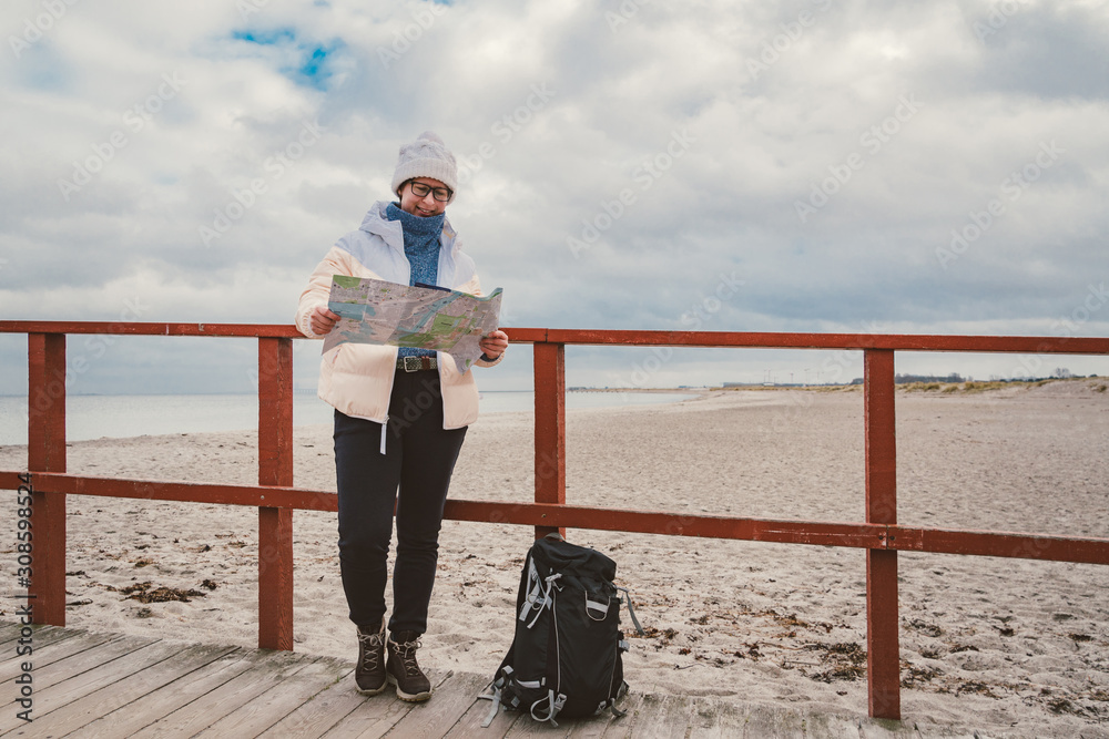 Caucasian woman in hat and jacket with backpack in winter sits on wooden pier on beach near North Sea. Denmark Copenhagen tourist uses paper map shore. Theme Travel and Navigation in Europe