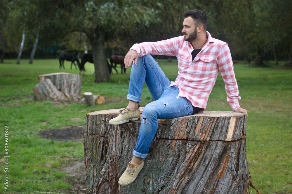 Fototapeta premium Young handsome guy Cowboy. farmer is sitting on a large stump on his ranch. Rural landscapes, countryside. Trees, field, farm. Stock photos for design.