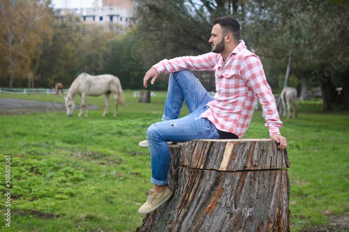 Young handsome guy Cowboy. farmer is sitting on a large stump on his ranch. Rural landscapes, countryside. Trees, field, farm. Stock photos for design. photo