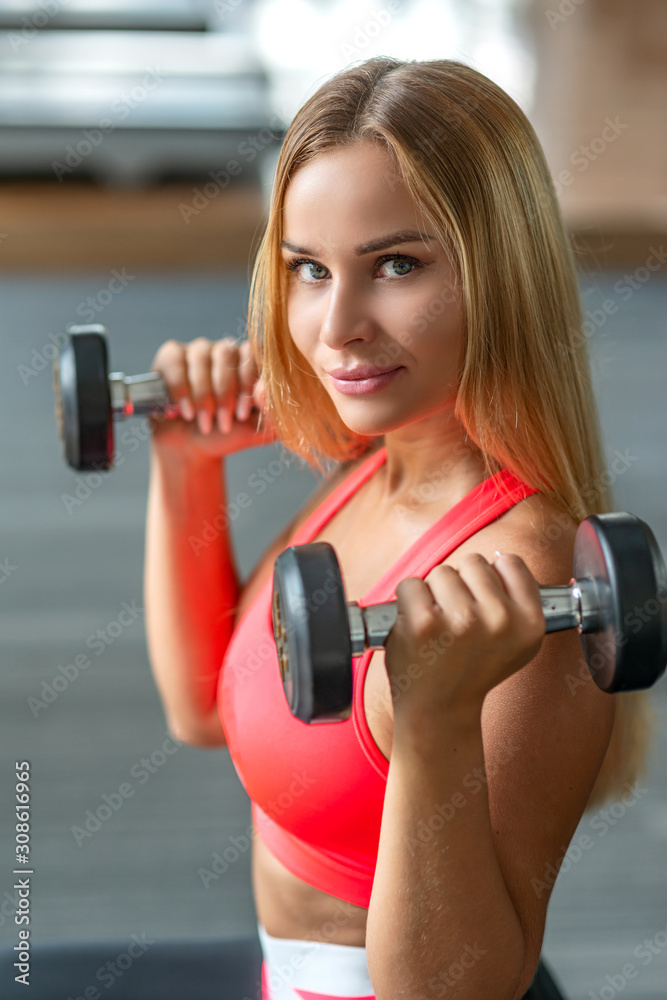 Fototapeta premium Portrait of sexy fit blonde in stylish sportswear doing the exercises with dumbbells in gym. Fitness woman in training. Sporty girl, workout concept. Vertical shot