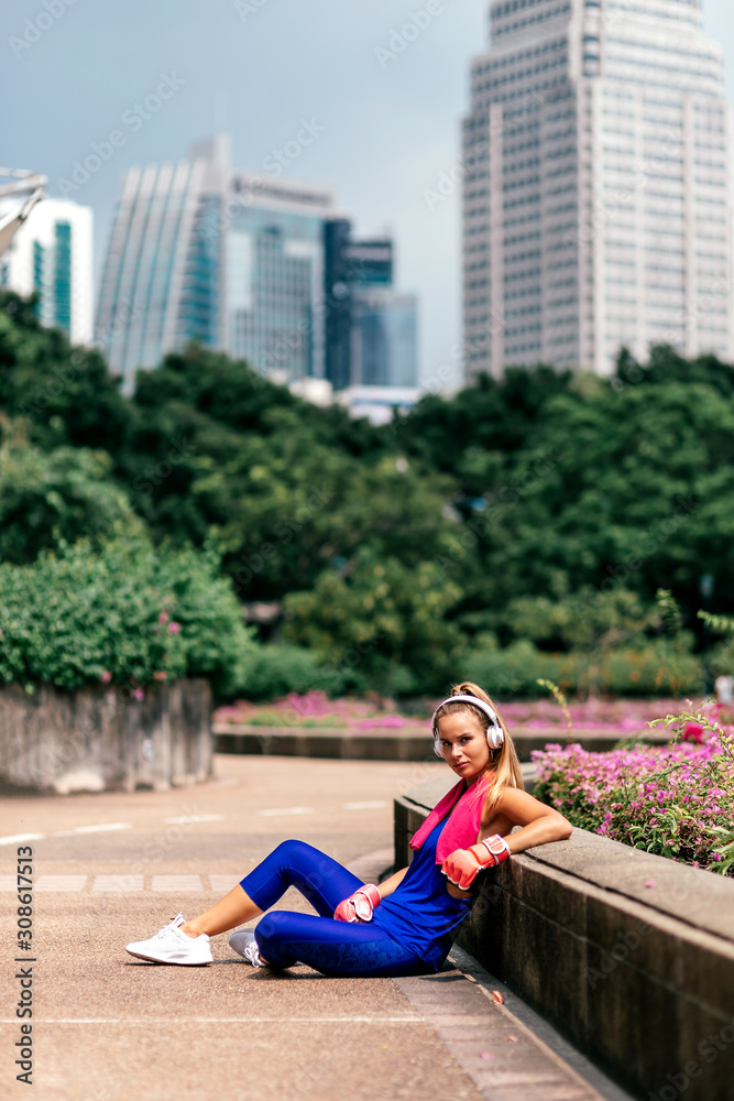 Full-length portrait of fit blonde in stylish bright sportswear sitting in city park after training. Fitness woman listening to music via headphones while relaxing. Sporty girl concept. Vertical shot