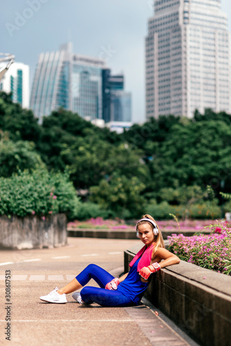 Full-length portrait of fit blonde in stylish bright sportswear sitting in city park after training. Fitness woman listening to music via headphones while relaxing. Sporty girl concept. Vertical shot