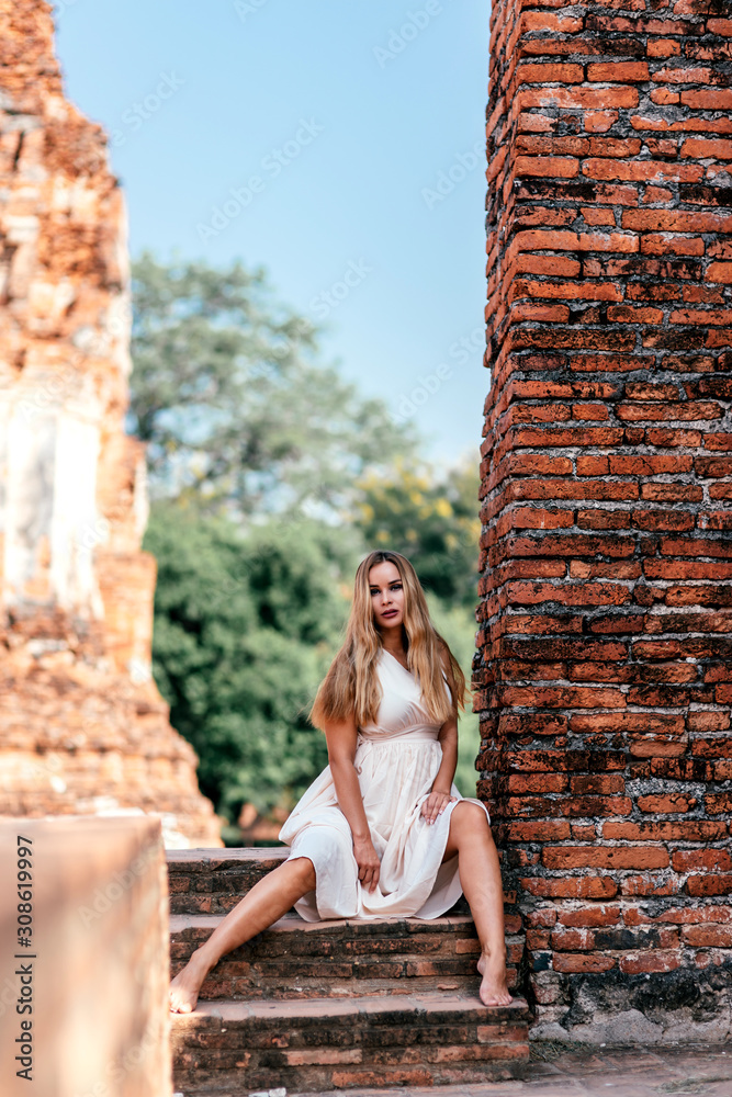 Portrait of attractive tourist girl on vacation looking at camera while having photoshoot in white dress near old ruins. Summer trip, tourism, adventures concept. Vertical shot