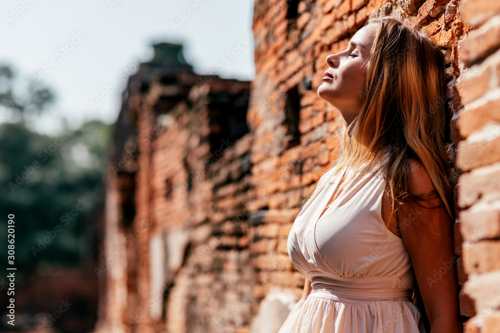 Portrait of beautiful blonde girl in white dress leaning on brick wall and looking up while posing outdoors. Summer vacation, adventures concept. Horizontal shot. Side view