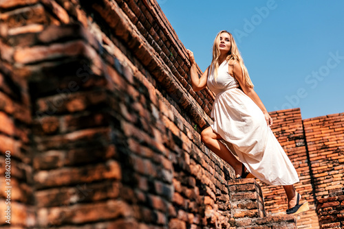 Full-length portrait of attractive tourist girl on vacation looking aside while having photoshoot in white dress near old ruins. Summer trip, tourism, adventures concept. Horizontal shot. Low angle © Adamov