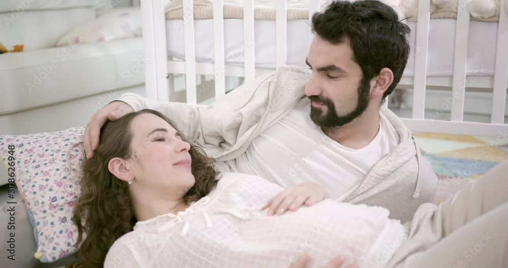Man and Pregnant Woman Sitting on Floor, Embracing and Laughing. Young Family Expecting a Baby. White Interior with Baby Cot on the background. Concept of Parenthood, Happiness and Tenderness Man and Pregnant Woman Sitting on Floor, Embracing and Laughing. Young Family Expecting a Baby. White Interior with Baby Cot on the background. Concept of Parenthood, Happiness and Tenderness