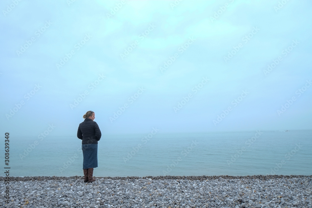 Woman wearing warm clothes and a purse looking out over a foggy sea from a pebble beach on the shore Woman wearing warm clothes and a purse looking out over a foggy sea from a pebble beach on the shore