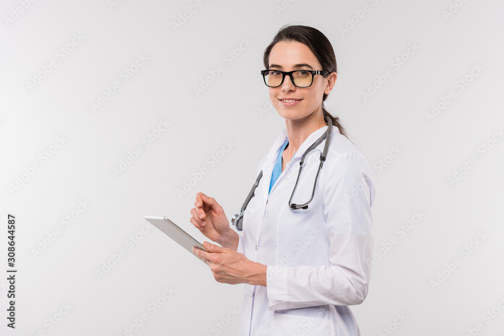 Young female clinician in whitecoat and eyeglasses using touchpad in isolation Young female clinician in whitecoat and eyeglasses using touchpad in isolation