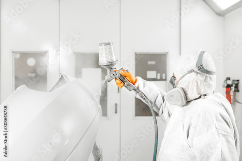 Automobile repairman painter in mask and protective workwear painting white car body in paint chamber. © Ilshat