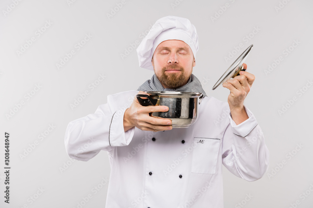 Young professional chef enjoying nice smell of soup or other cooked meal in pan
