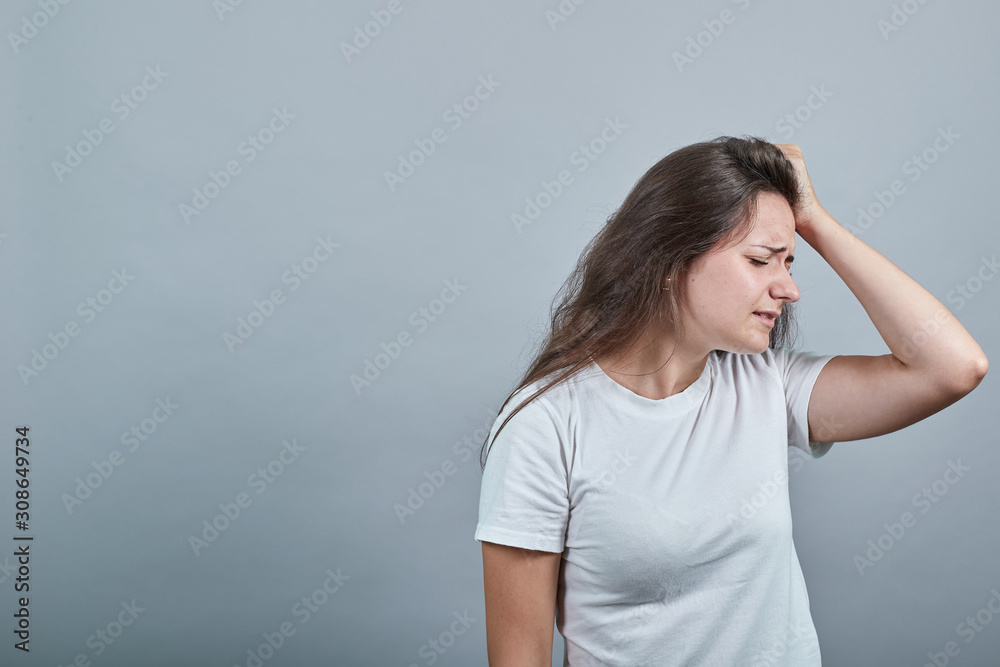 Young lady in white t-shirt over isolated wall holds her palm on her head, she has headache. Girl looks sad, unhappily and worried