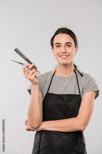 Young female hairdresser holding hairbrush and scissors and looking at you
