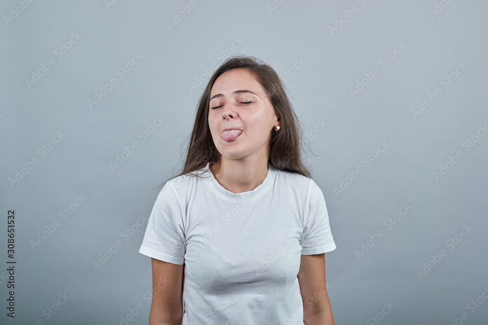 Teen in white t-shirt shows her tongue in jest. Gils looks childish and windy. Behind her is gray wall. Hair is flowing down. Teen in white t-shirt shows her tongue in jest. Gils looks childish and windy. Behind her is gray wall. Hair is flowing down.