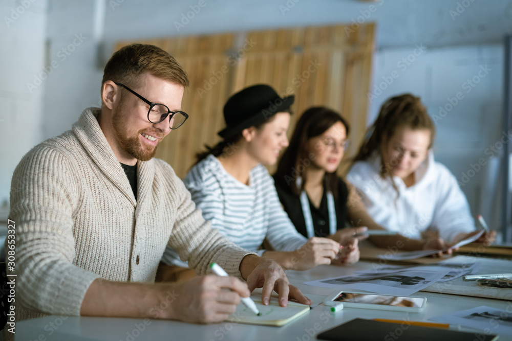 Creative fashion designer drawing sketch in copybook on background of colleagues Creative fashion designer drawing sketch in copybook on background of colleagues