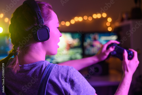 Gamer girl with a joystick plays on a console or computer using a gamepad, controller, joystick. Against the background of three monitors, with creative light © Anton