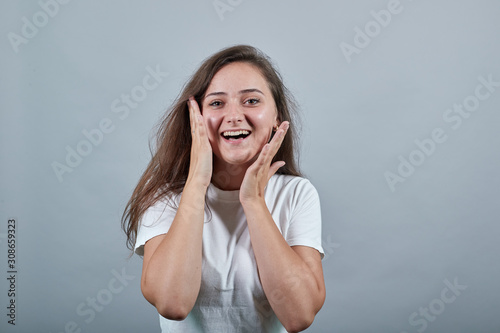 Young teenager student girl with long chestnut hair over isolated gray wall laughing and holding her hands near cheeks. She looks happy, joyfull and cheerfull.