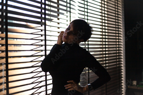 Silhouette of a beautiful 45s years old woman in black style clothes on a background of jalousie indoors at the house.