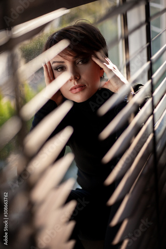 Portrait of beautiful woman in black style clothes sitting on the window sill behind the jalousie indoors at the house. The sun's beam on eye of a peeking model. The striped shade on the face.