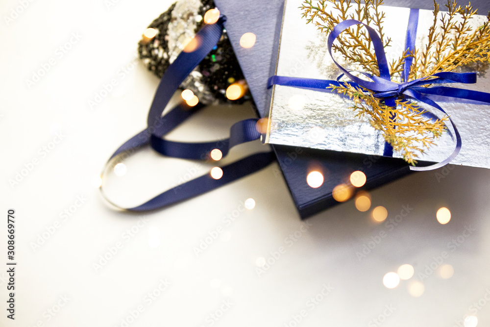 Christmas composition - blue paper bag with golden box gifts with blue bow ribbon, Xmas bauble, balls on white background with lights. Creative greeting card in trend colors, top view, copy space