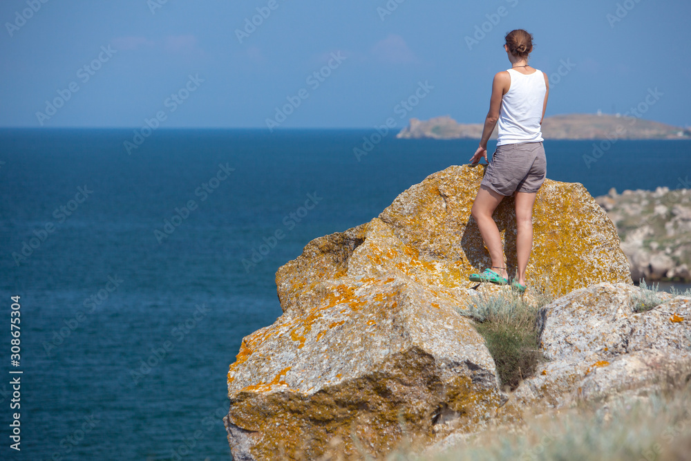 Young blond tourist woman standing on natural rocks by the sea at sunset and looking aside. Mediterranean region, Turkey