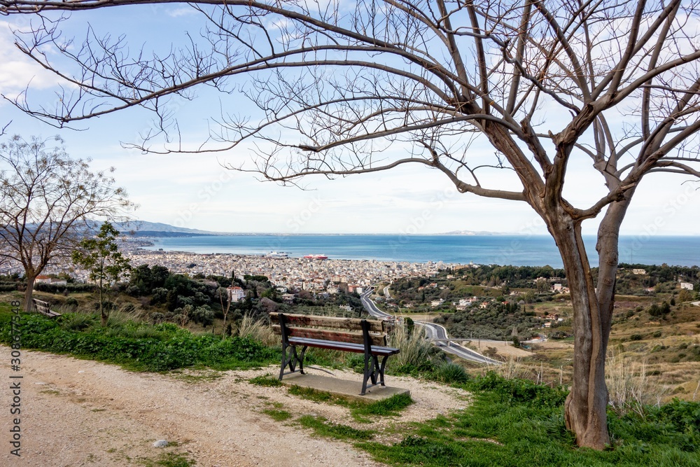 The view from lookout above the Patras city in Greece with a tree and a bench to relax and admire the beautiful cityscape The view from lookout above the Patras city in Greece with a tree and a bench to relax and admire the beautiful cityscape