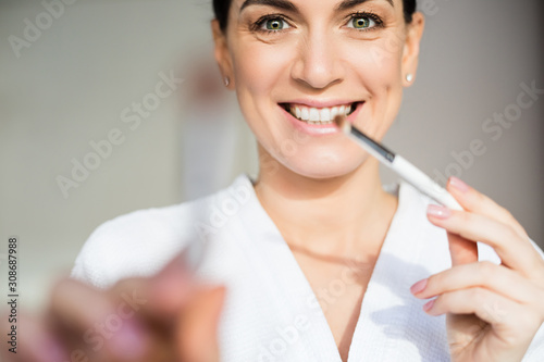 Portrait of beautiful brunette smiling middle age woman wearing white bathrobe looking into the camera and holding brushes in hands