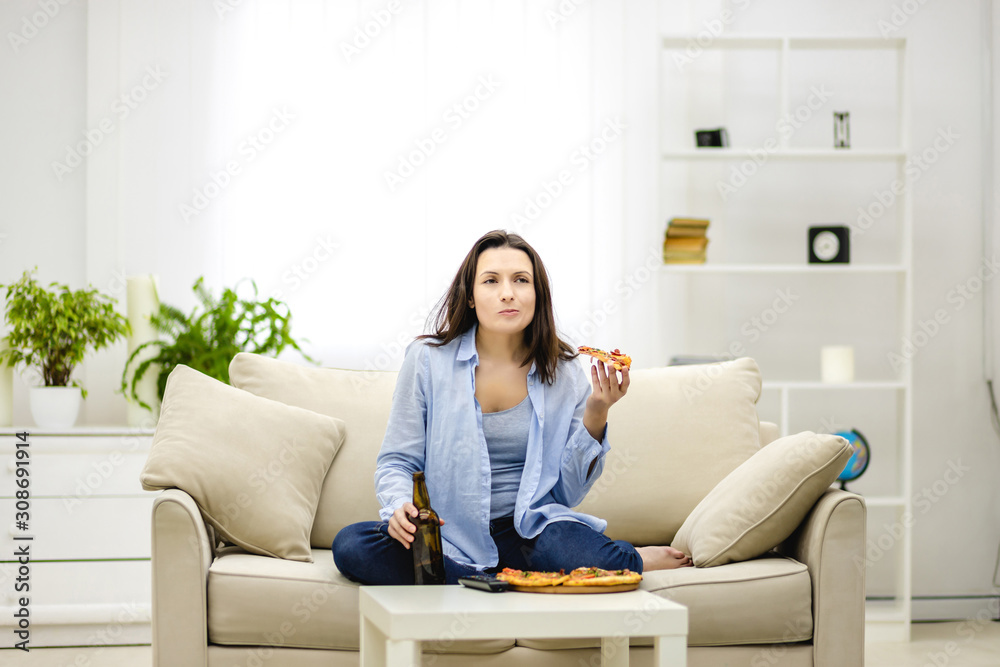 Hungry woman chews, holding a bottle of beer and a slice of pizza hands. Isolated woman on blurred light background. Copy space. Hungry woman chews, holding a bottle of beer and a slice of pizza hands. Isolated woman on blurred light background. Copy space.