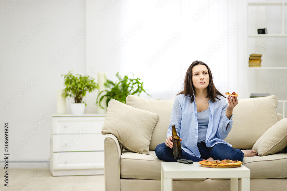 Brunette woman chews, holding a bottle of beer and a slice of pizza hands. Isolated woman on blurred light background. Copy space. Brunette woman chews, holding a bottle of beer and a slice of pizza hands. Isolated woman on blurred light background. Copy space.