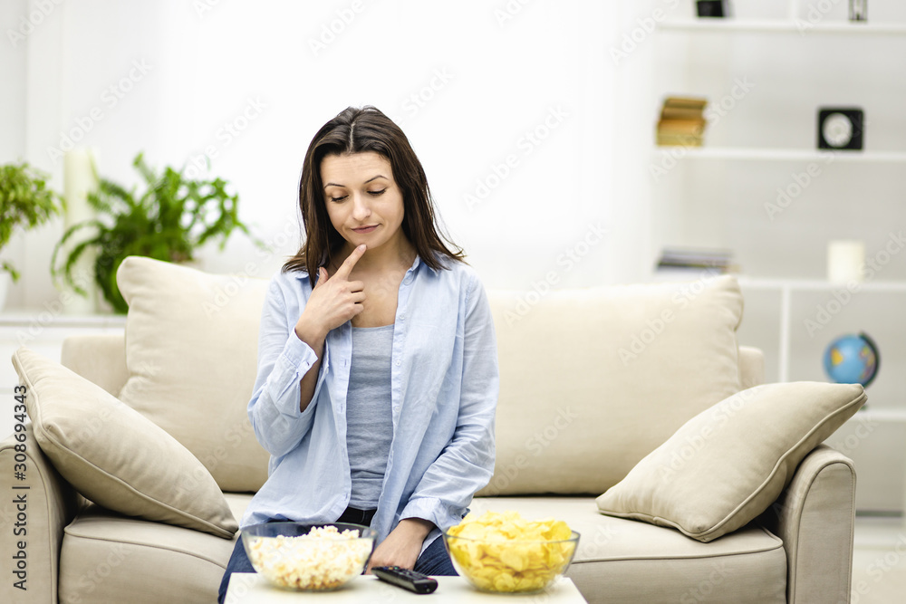 Puzzled brunette woman is thinking which snack to choose, isolated, sitting on the couch at home. Puzzled brunette woman is thinking which snack to choose, isolated, sitting on the couch at home.