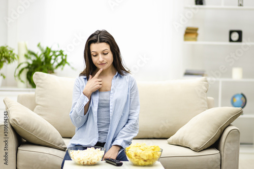 Puzzled brunette woman is thinking which snack to choose, isolated, sitting on the couch at home.