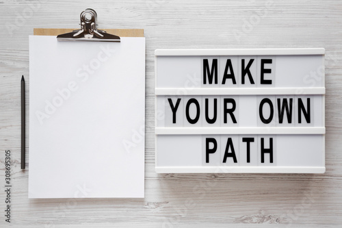 Fototapeta Naklejka Na Ścianę i Meble -  'Make your own path' words on a lightbox, clipboard with blank sheet of paper on a white wooden background, top view. Overhead, from above, flat lay. Copy space.