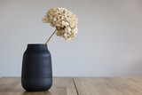 A black ceramic vase with a dried hydrangea flower stands on a wooden surface.