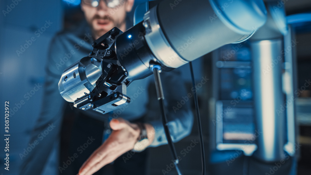 Close Up of a Futuristic Robotic Arm Moving a Metal Detail. Team of Engineers Discuss This Technologically Advanced Process. They are in a High Tech Research Laboratory with Modern Equipment. Close Up of a Futuristic Robotic Arm Moving a Metal Detail. Team of Engineers Discuss This Technologically Advanced Process. They are in a High Tech Research Laboratory with Modern Equipment.