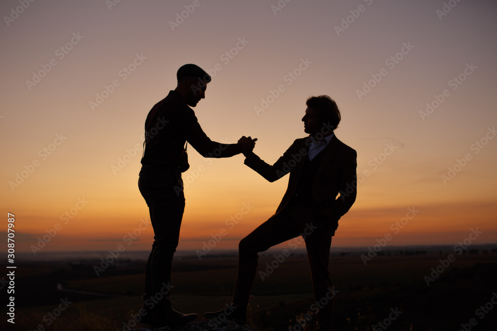 Silhouette of two men in sunset sky (orange and pink sky) on rock hill. Two gentlemen are taking, looking and agree each other, then shake hands. Agreement and cooperation concept