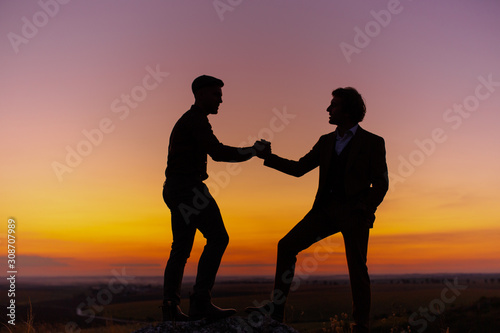 Silhouette of two men in sunset sky (orange and pink sky) on rock hill. Two gentlemen are taking, looking and agree each other, then shake hands. Agreement and cooperation concept
