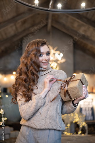 Beautiful woman (girl) dressed in a warm fashion long sleeve sweater among Christmas lights with a gift box with a bow