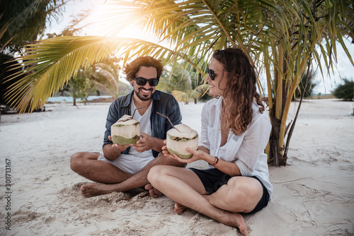 A beautiful young couple joyfully sits on white sand under a palm tree with coconuts in their hands on the seashore under a green palm tree. Honeymoon Travel and Vacation on the Tropical Ocean.