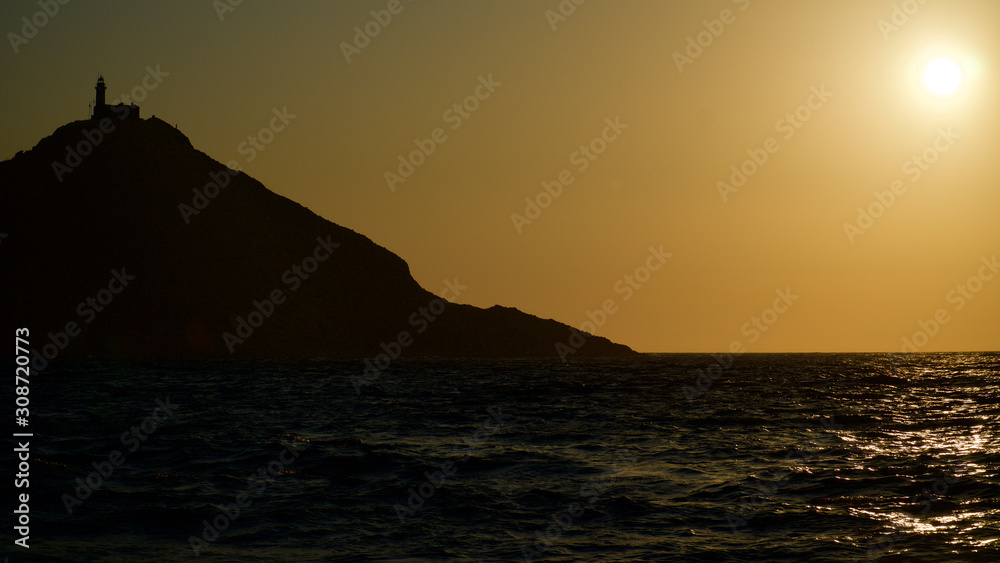 Spectacular sunset with endless horizon and sparkling waves. Lighthouse and rocky hills in the background.